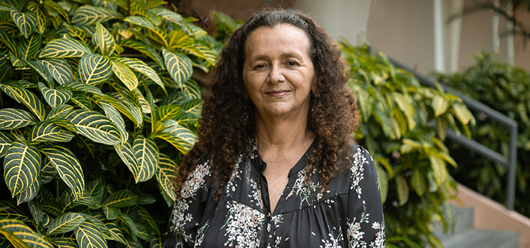 #paratodosverem: Fotografia mostra, em primeiro plano, a professora Ana Cleide Moreira vestindo camisa preta com estampas brancas. Ela tem cabelos encaracolados abaixo dos ombros e sorri para nós. No fundo, folhagem natural verde e amarela.